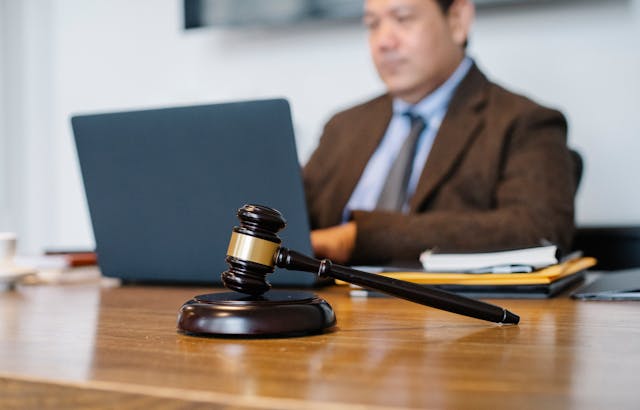 A gavel sitting on a desk with a person using a laptop in the background