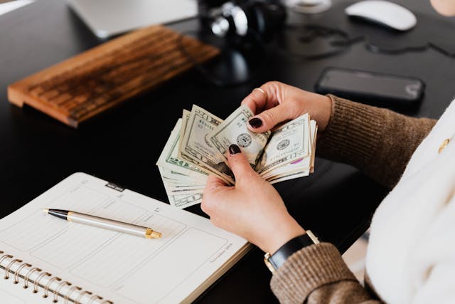 A person sitting at a desk counting a stack of money, with a notebook and pen in the background.