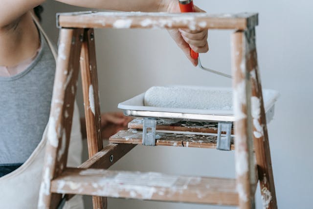 Person rolling a paint roller in a paint tray attached to a ladder