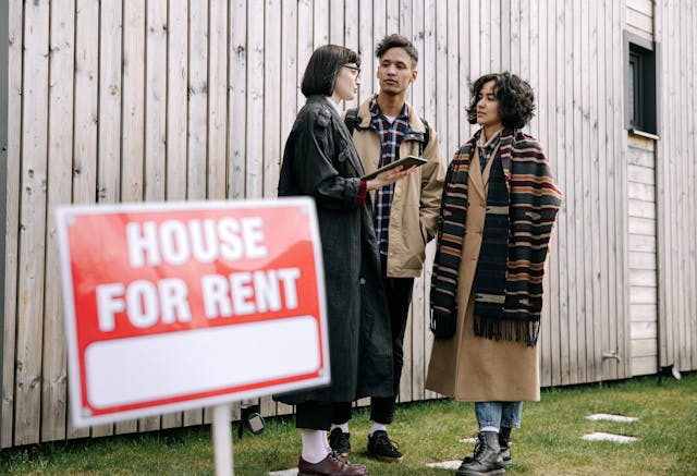 Three people standing outside behind a house for rent sign