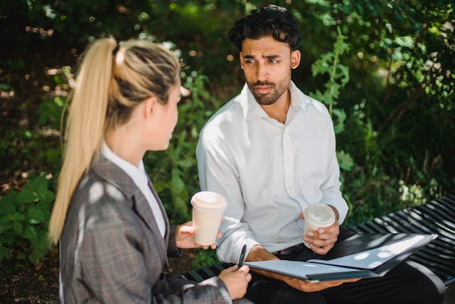 Two people sitting outdoors on a bench, holding coffee cups, discussing documents in a folder.